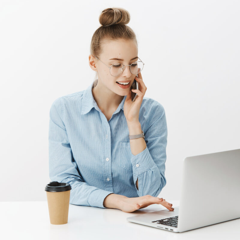 Portrait of charming hardworking female freelancer working with laptop looking at screen, checking order as talking via smartphone with customer, wearing glasses and blue shirt, drinking coffee.