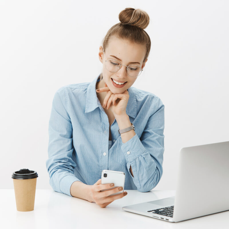Good-looking friendly and cheerful young female office worker in shirt and glasses watching video in smartphone and smiling as having rest of work sitting near laptop and drinking coffee. Technology, finance and business concept