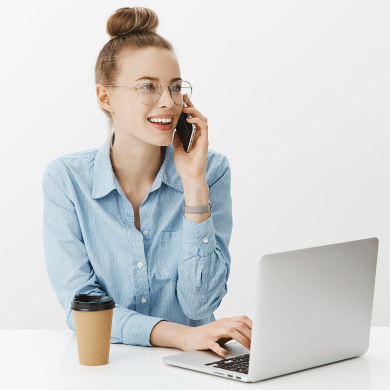 Girl having talk with customer via smartphone as sitting in office working with laptop, drinking coffee, leaning on table, smiling polite and pleasant as leaning on table with elbow over gray wall.
